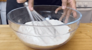 Person whisking flour in a glass bowl on a wooden surface, preparing ingredients for baking.